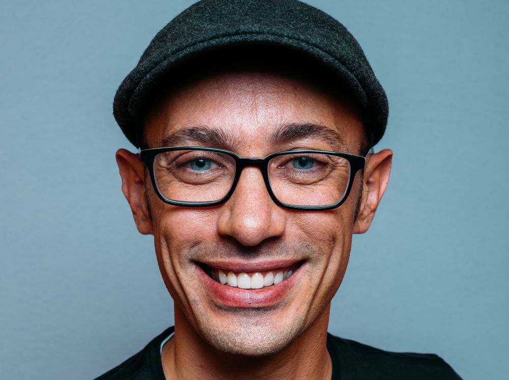A closeup headshot of Tobi L&uuml;tke smiling into the camera sporting black-framed glasses and a flat cap. 