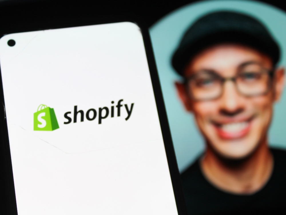 The Shopify logo displayed on a smartphone screen. Shopify CEO Tobi L&uuml;tke&rsquo;s Instagram photo can be seen in the background out of focus: a middle-aged man wearing glasses and a cloth cap and smiling broadly.