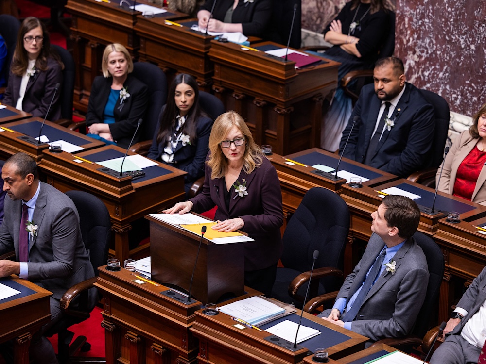 A middle-aged woman with light skin and long blond hair wearing a suit stands at her desk in B.C.&rsquo;s legislature chamber. Other MLAs wearing suits and other formal business attire sit around her.