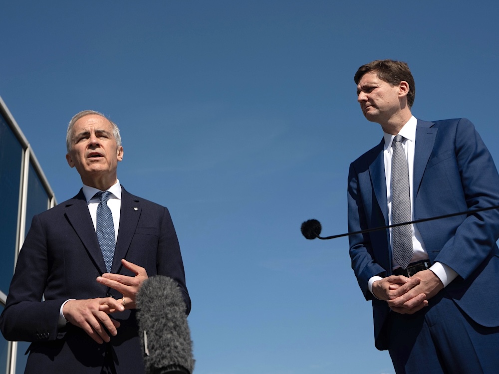 Two suit-wearing men stand before a blue sky. One speaks into a microphone positioned below and in front of him.
