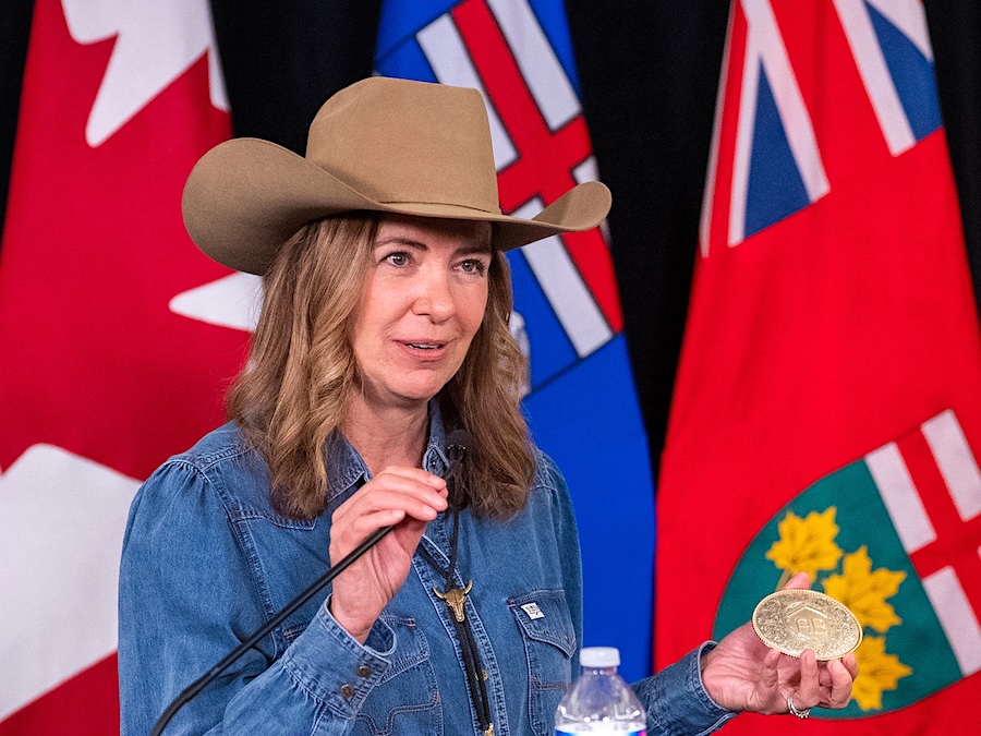 Danielle Smith, a 55-year-old light-skinned woman, stands at a podium. She wears a denim jacket and cowboy hat.