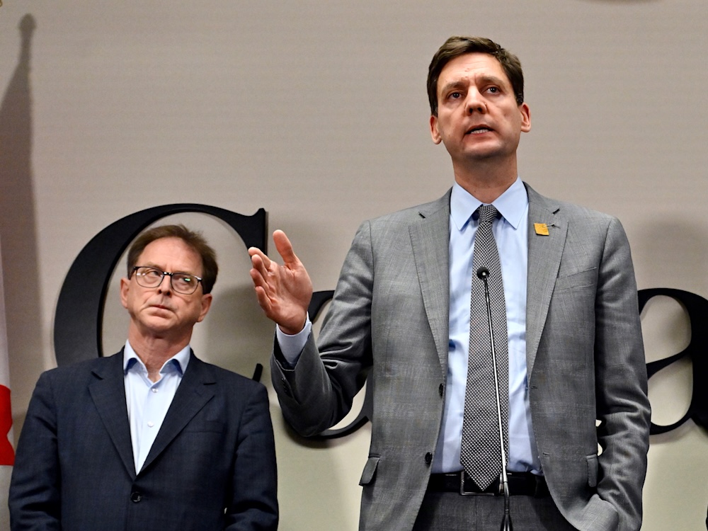 A tall middle-aged white man, B.C. Premier David Eby, stands and gestures in an indoor venue with letters belonging to the word &lsquo;Canada&rsquo; behind him. Small and to his right is a bespectacled middle-aged white man, Minister Adrian Dix.