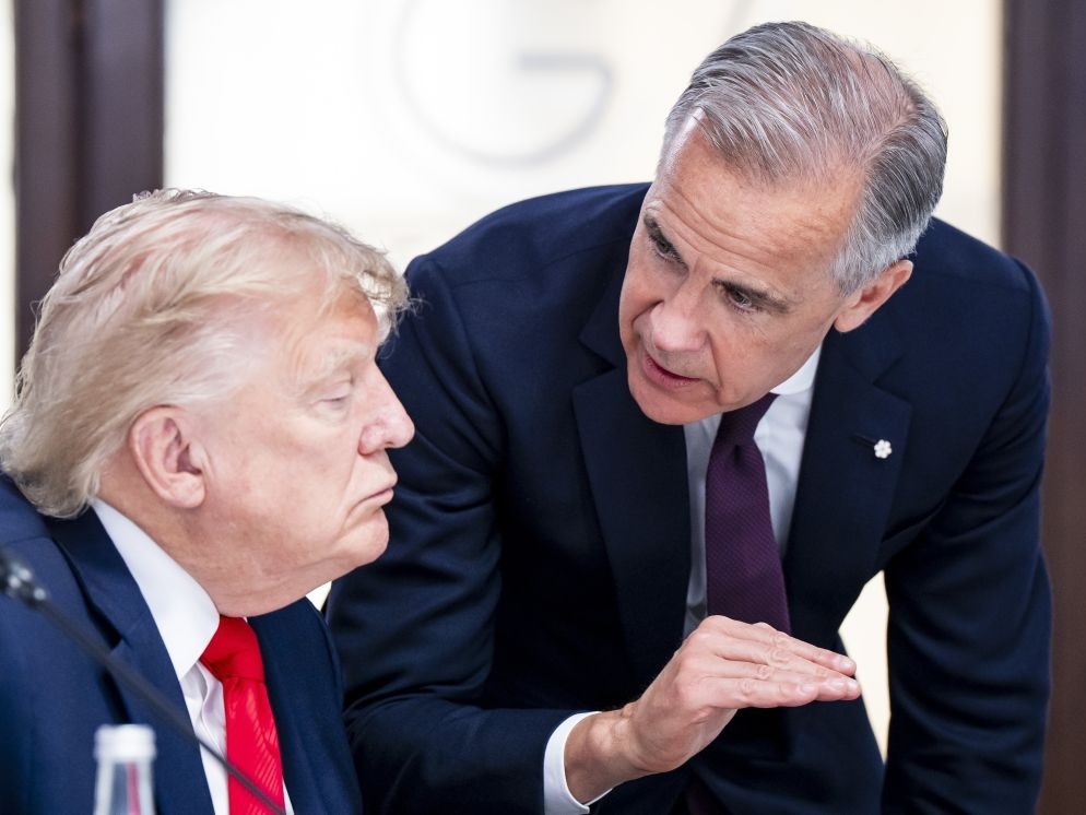 A grey-haired white middle-aged man, Prime Minister Mark Carney, stands and leans over making an emphatic gesture with his hand as a jowly white elderly man, President Donald Trump, sits at a table with his eyes closed and lips pursed.