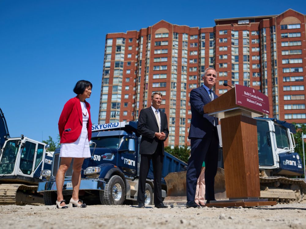 Mark Carney stands at a podium at a construction site, in front of a large, multi-storey apartment building.