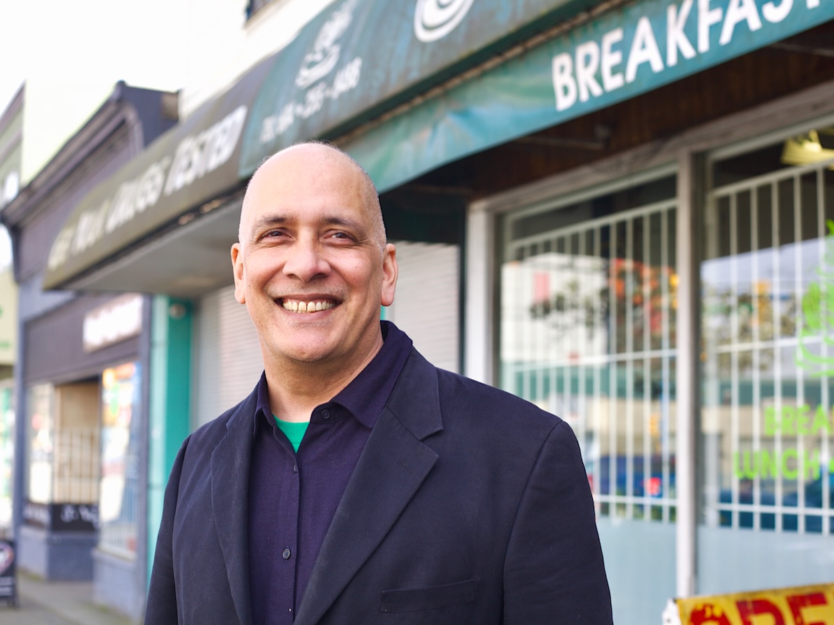 A middle-aged man stands on a city street with business awnings and storefronts in the background. He is bald, has a broad, friendly smile and wears a navy blue blazer and navy blue button-down shirt.