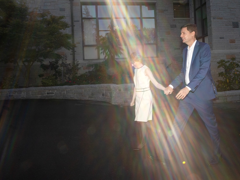 A tall light-skinned man in a blue suit and a woman in a white dress hold hands as they walk in a bright sunbeam in front of a stone building.