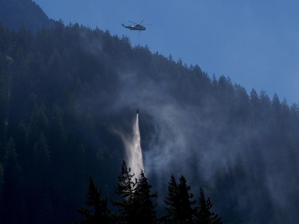 A helicopter flies over a forested mountain range, pouring buckets of water on it against a blue sky.