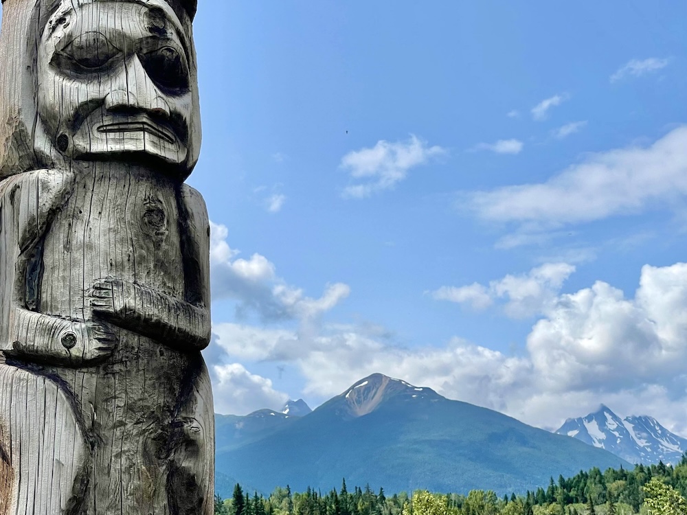 A totem pole to the left of the frame stands against a clear sky and a blue mountain range.