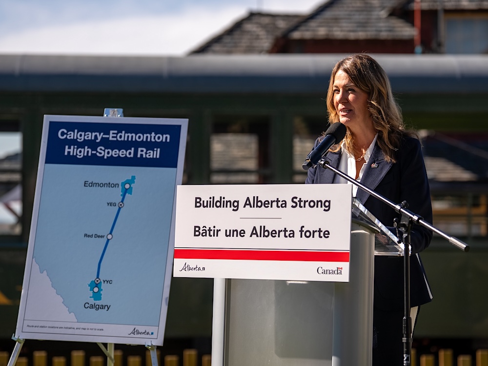 Danielle Smith stands outside at a podium, with a map showing a train route between Edmonton and Calgary.