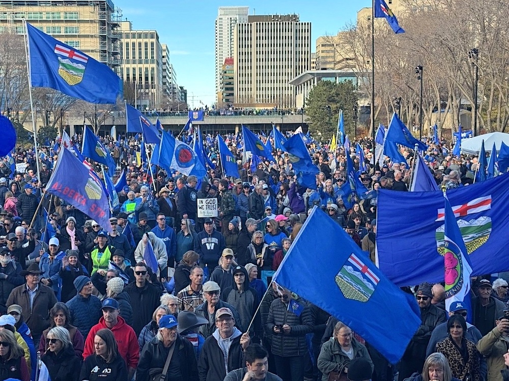 A large crowd of people gathers in an urban public area, with many holding blue Alberta flags.