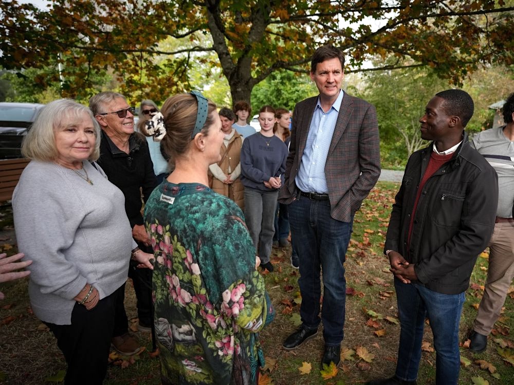 A tall light-skinned middle-aged man in a sports coat, David Eby, speaks under a tree. Around him are a dark-skinned young man, George Anderson, and two light-skinned women of middle age.