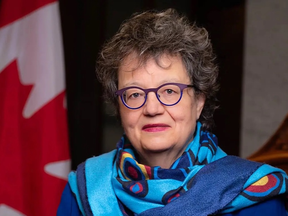 Paula Simons has light skin tone and short wavy brown hair with highlights. She is wearing a blue patterned scarf and purple and blue rimmed eyeglasses. A Canadian flag is to the left of the frame.
