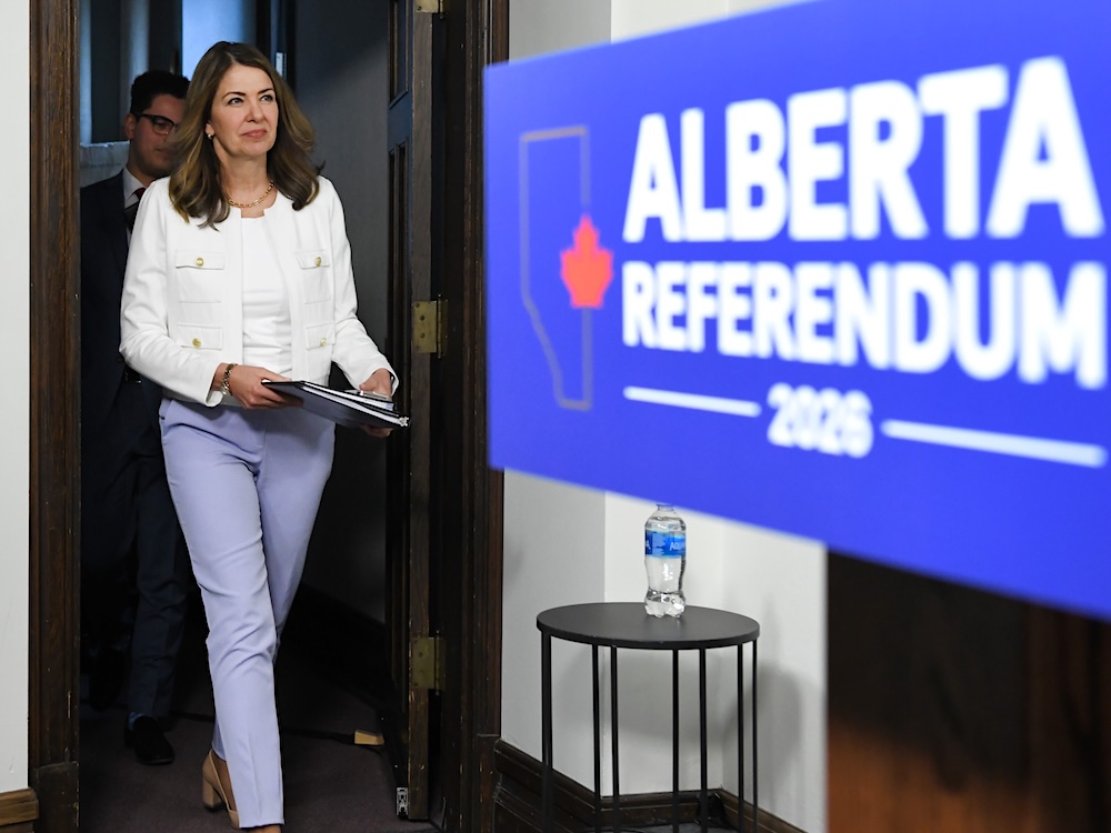 Danielle Smith walks onto a stage with a blue &lsquo;Alberta Referendum&rsquo; sign. She wears lilac pants and a white shirt and jacket.