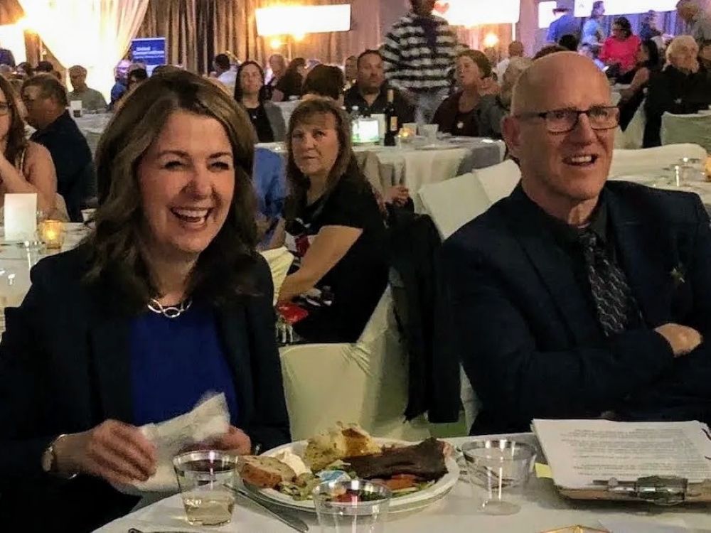 A 55-year-old light-skinned woman with long dark hair sits at a banquet table, smiling, beside a 60-ish bald man wearing a suit.
