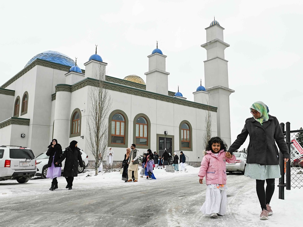 A woman in a green headscarf and dark winter coat holds the hand of a young girl with a pink winter jacket as they walk on a snowy driveway. They and others in winter clothing are leaving a white mosque with bright blue rounded accents on the roof.