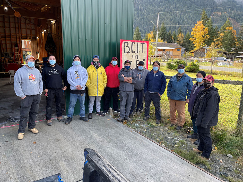 Eleven people wearing masks stand in a semi-circle. They are outdoors and outside a building with a large, open garage door. In the background, there are roads, a few homes, and a forested mountain.