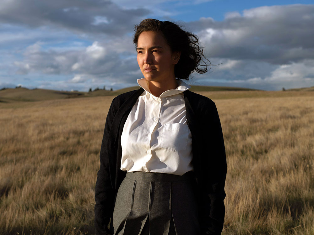 A woman wearing a white button-up blouse and black cardigan stands in a prairie field. The sky is crisp blue with scattered clouds, the grass is dry. A light breeze blows through the woman's hair as she gazes deeply into the distance.
