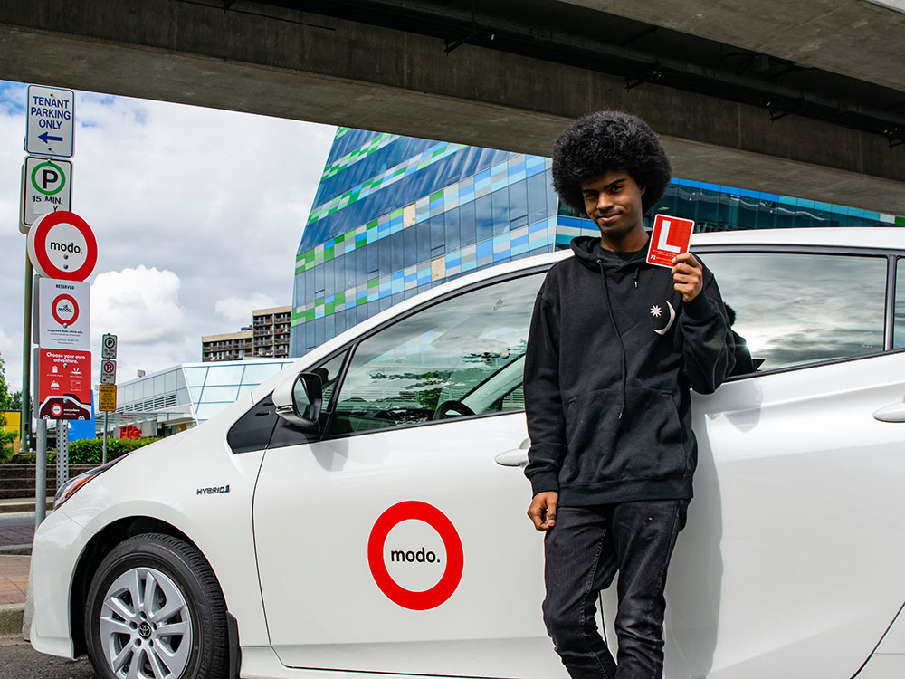 A youth leans on a Modo-branded car holding an “L” permit magnet. They are parked under the SkyTrain, near a building with colourful glass windows.