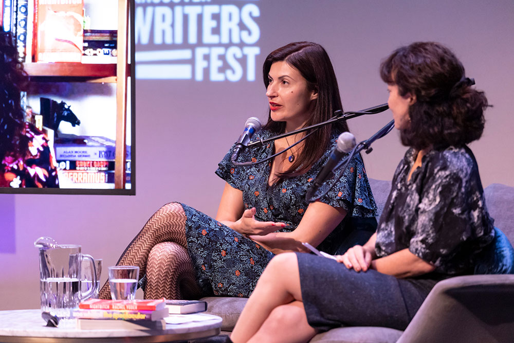 Two women sit on a couch as part of a discussion panel. A screen with other contributors, participating digitally, is visible on the left pane.