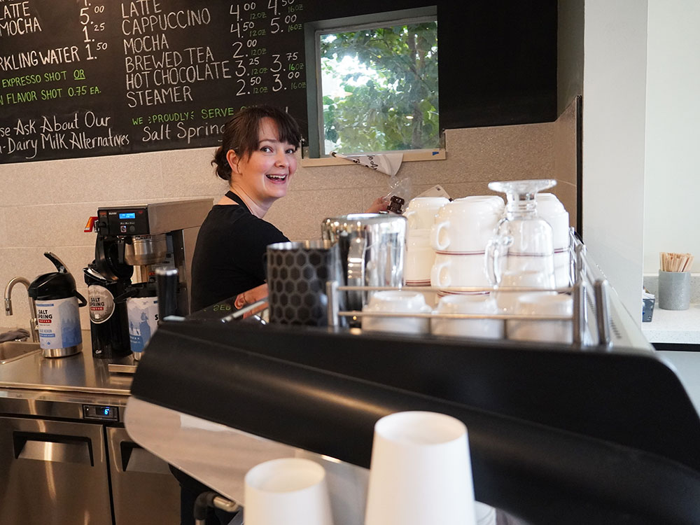 Corrie Corfield stands behind a commercial cappuccino machine stacked with ceramic and glass cups, turned to the camera with a smile, looking busy. A large chalk board café menu hangs on the wall behind her.