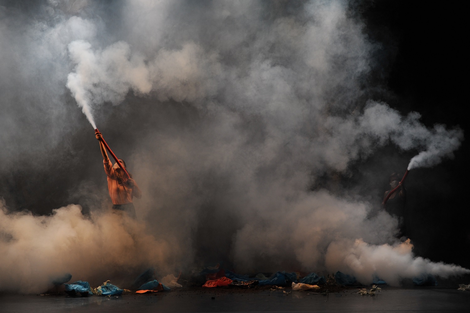 Two masked people on a black stage hold red tubes. Smoke is emerging from the tubes, so much that it is difficult to see. There appear to be small mounds of fabric on the ground all over the stage.