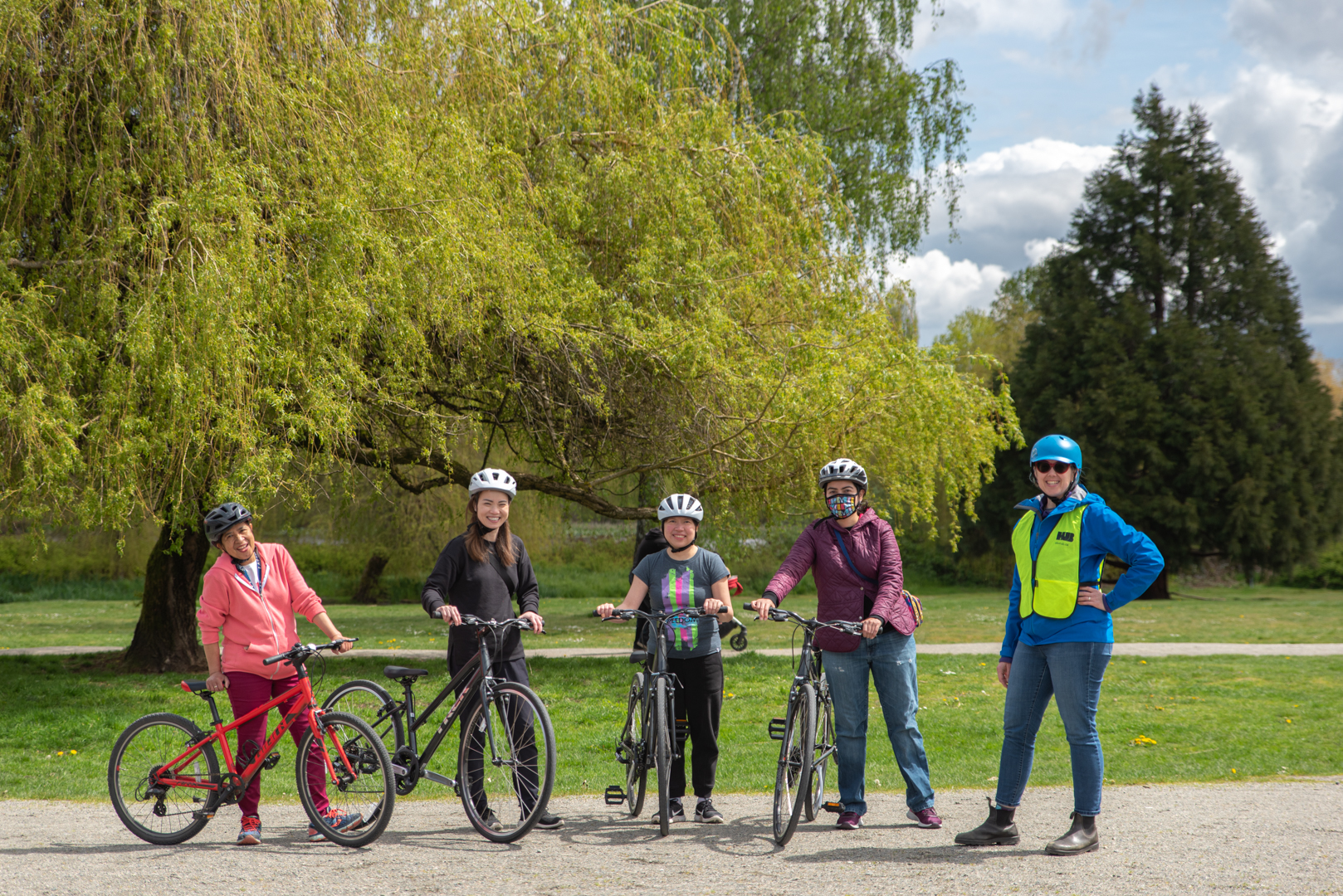 Four women stand side by side with their bikes, wearing helmets. A fifth women, without a bike, stands to their right. She is wearing a yellow safety vest.