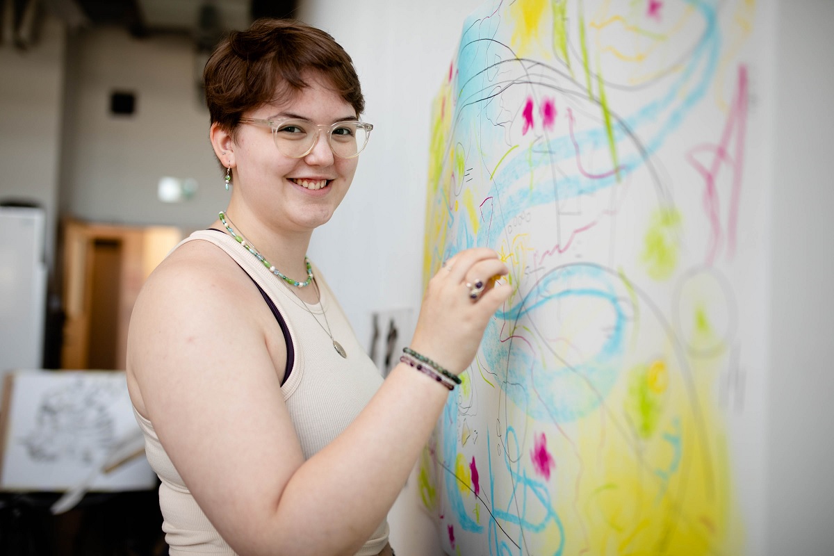 A young person wearing glasses is smiling at the camera, half turned, facing an easel and canvas.