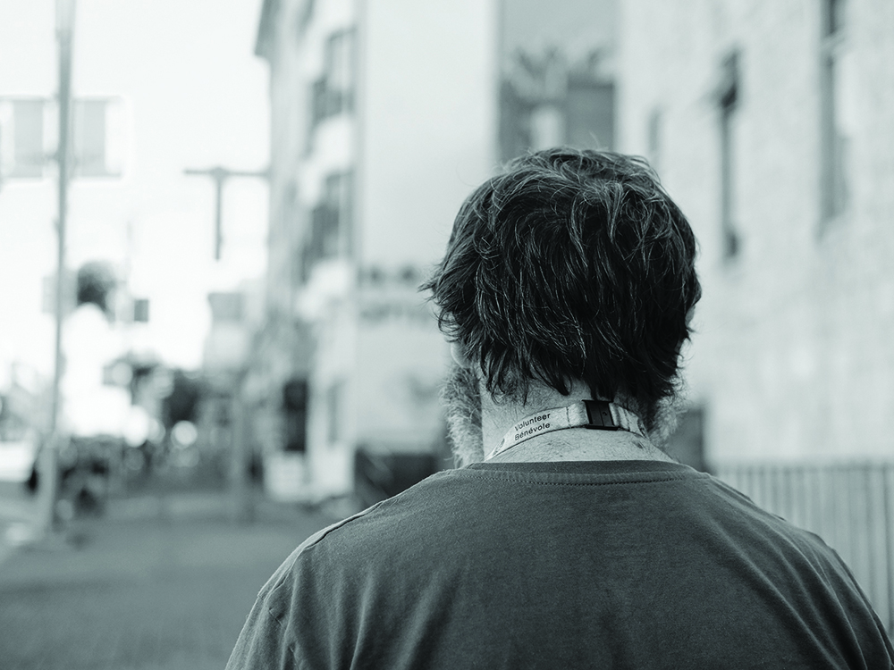 A photograph shows a man from behind. He is standing on a street in Vancouver, wearing a lanyard that says, “Volunteer / Bénévole.”