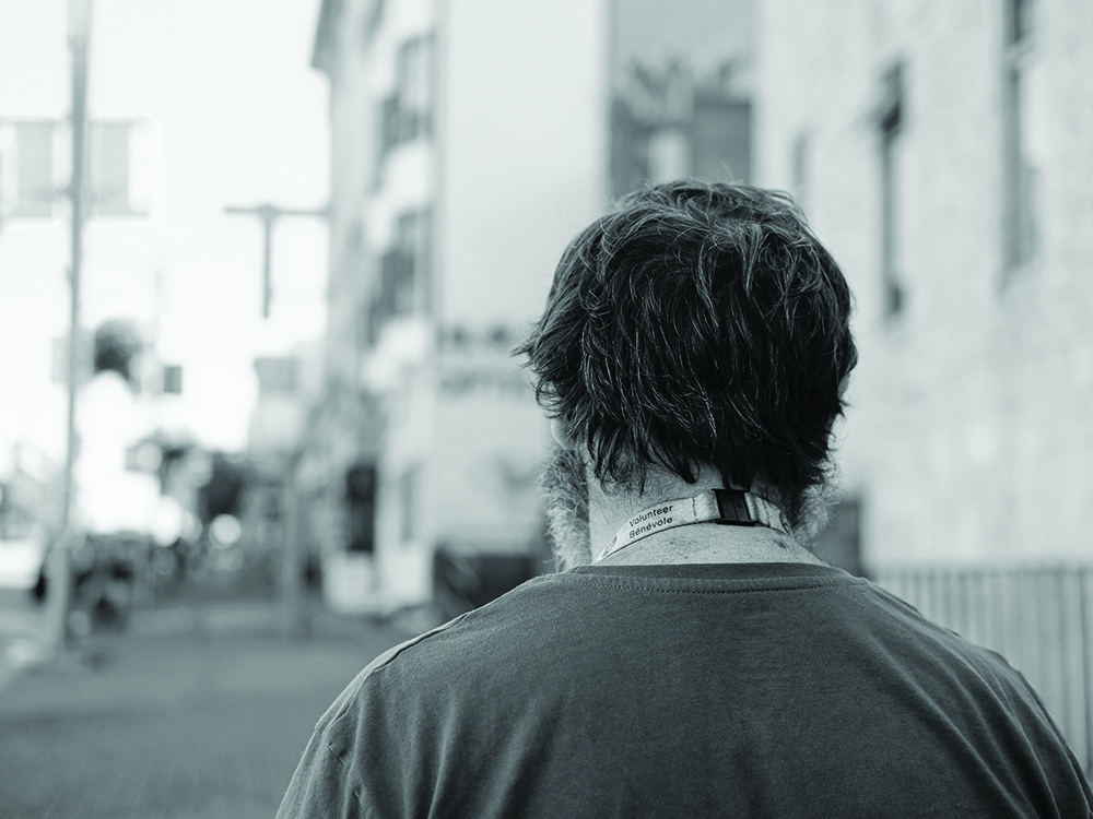 A photograph shows a man from behind. He is standing on a street in Vancouver, wearing a lanyard that says, &ldquo;Volunteer / B&eacute;n&eacute;vole.&rdquo;