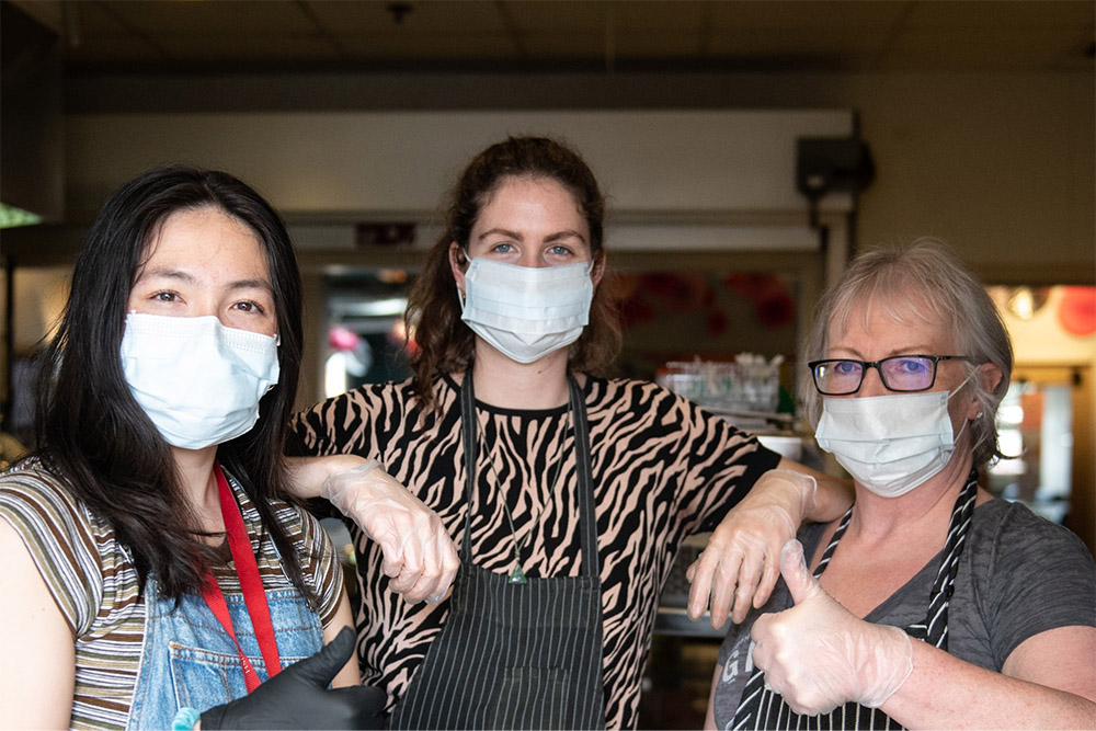 Three people stand together at the WISH Drop-In Centre Society wearing aprons. They are looking at the camera. One person is giving a thumbs-up. 