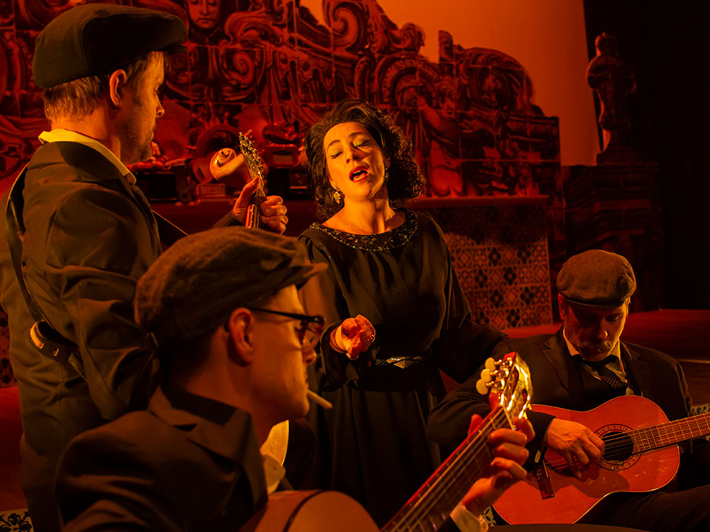 Three guitarists gather around a woman who is singing in a dark, red-tinged and old-style room. 
