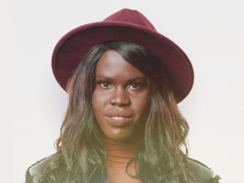 A closeup of a woman, Adhel Arop, looking directly at the camera. She is wearing an orange turtleneck and burgundy bucket hat.