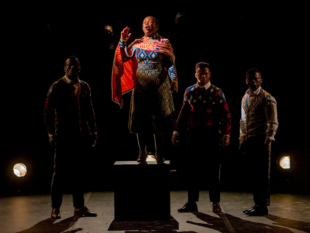 A Black woman in a colourful patterned dress and shawl stands on a box while singing. Three Black men in colourful patterned sweaters and black pants stand behind her in partial shadows.