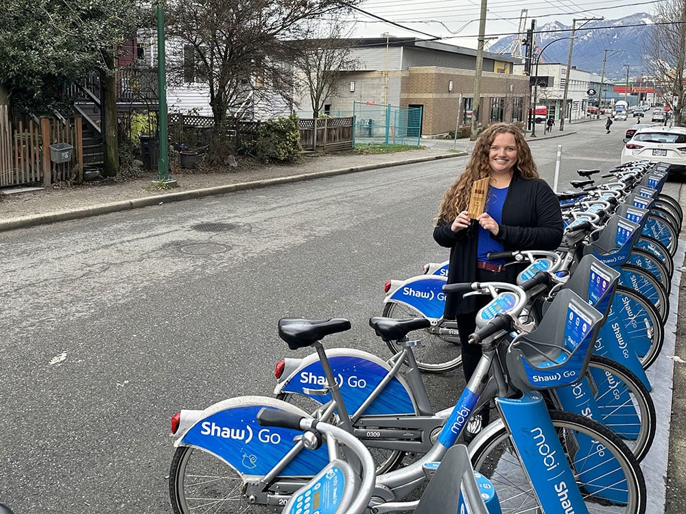 A woman with long curly hair stands holds a wooden award next to a row of Mobi bikes parked along a Vancouver street.