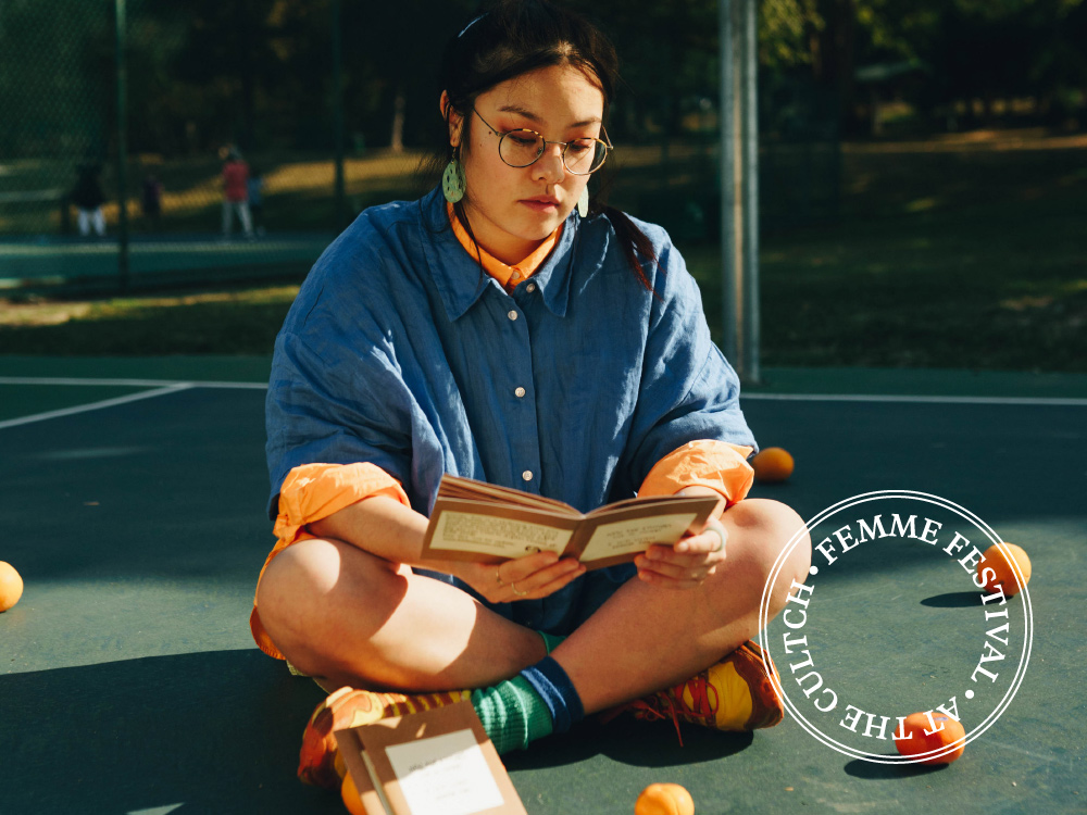 A person sits cross-legged on a tennis court reading a book, surrounded by oranges. 