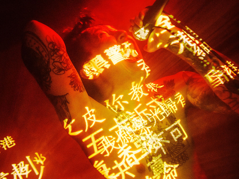 A man shrouded in red light and superimposed by Chinese characters looks up as if to be shouting from a mic. He is shirtless and has multiple tattoos on his arms and torso.