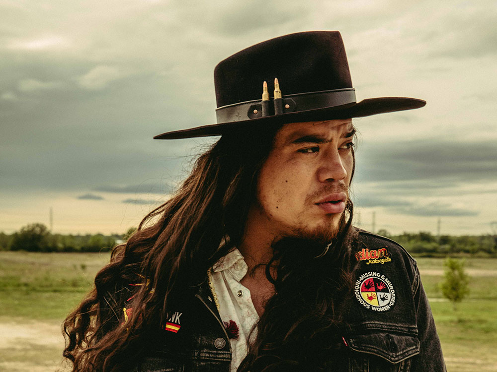 A photo of a man with long hair in a jean jacket stands in front of a prairie landscape, looking to the side.