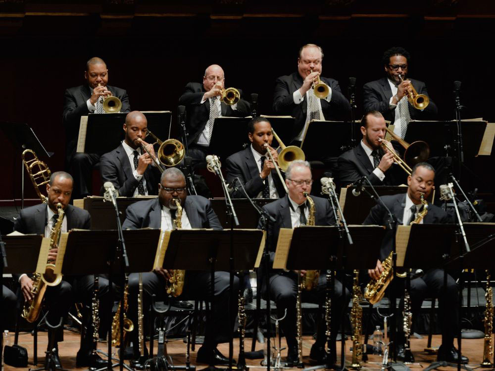A brass band orchestra of men in suits play in three rows, with music stands in front of them.