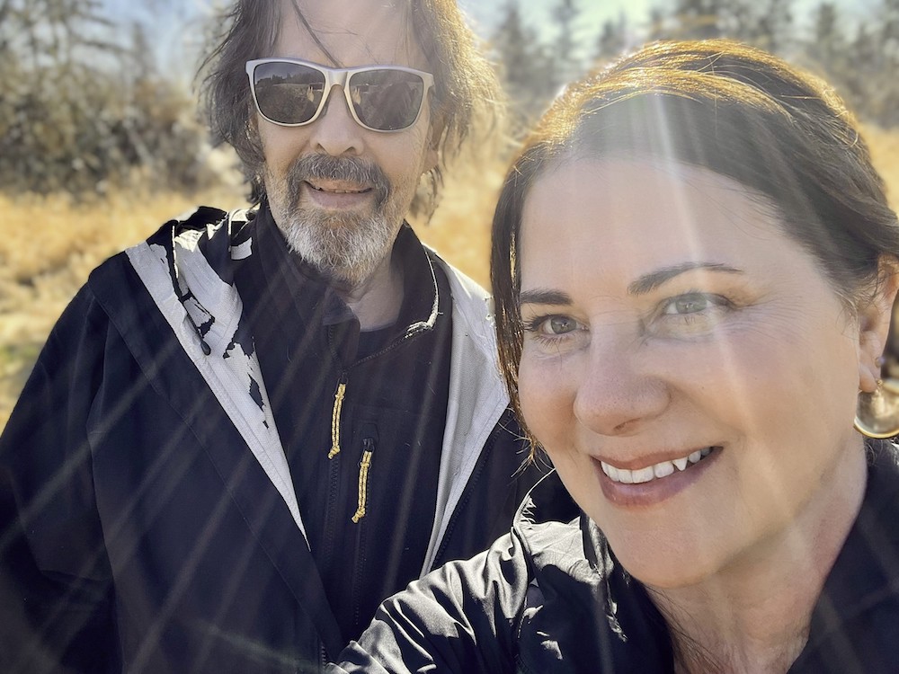 A selfie of a woman and man with rays of sun visible over their faces. The woman has brown hair and eyes and is smiling. The man has greyish hair and goatee and is wearing sunglasses.
