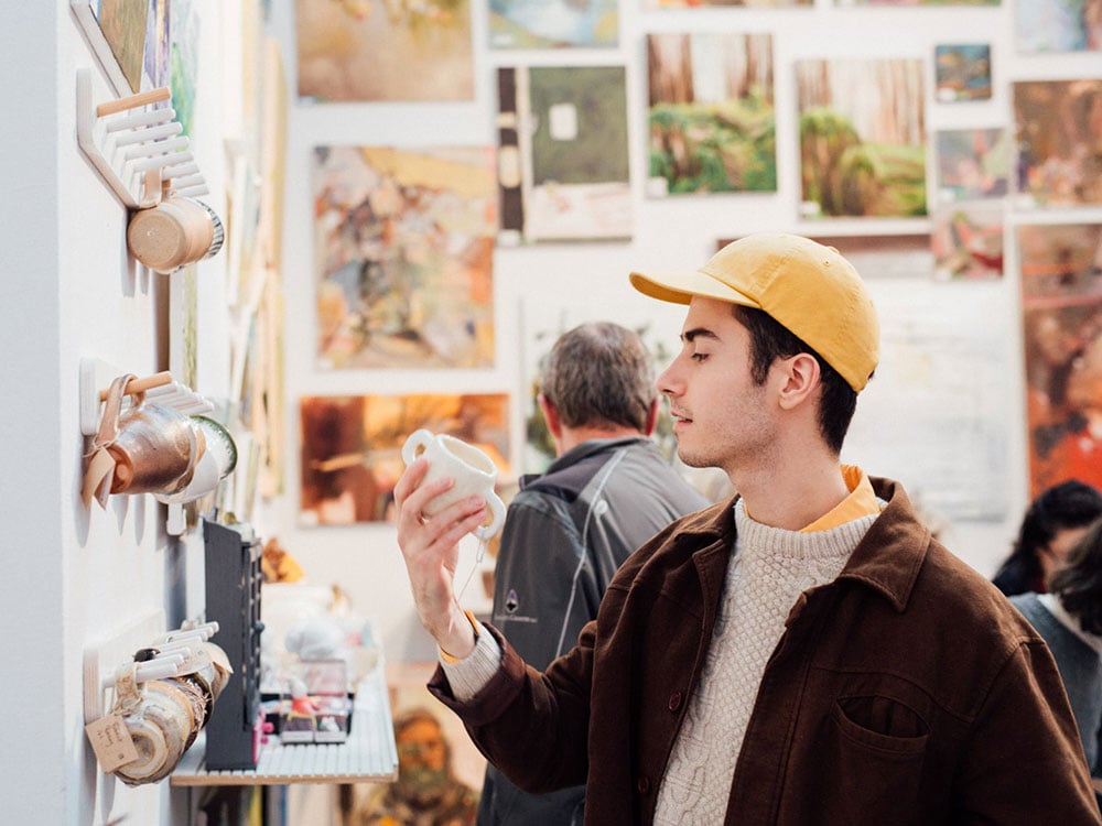 A person in a creamsicle orange baseball hat, a beige-knitted wool sweater and brown jacket holds and looks into a handmade white ceramic mug. Mugs hang on the wall in front of them, and a wall of tiled art hangs in the background.