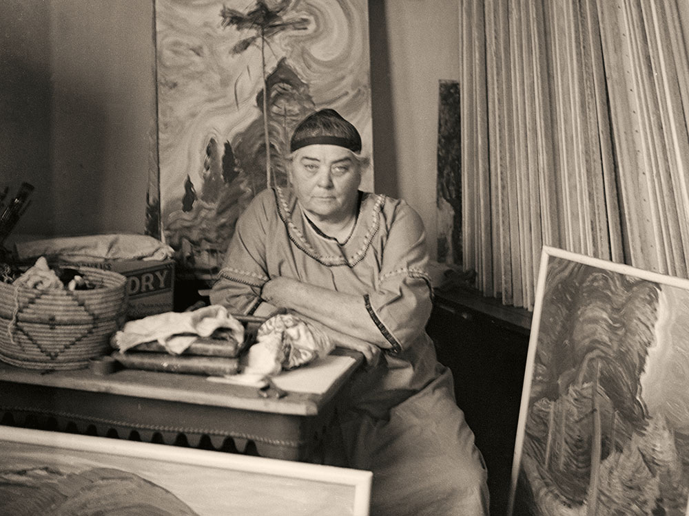 A middle-aged woman, Emily Carr, sits in a studio. She is leaning on a desk, staring directly at the camera, in a black cap and smock. A painting is propped on an easel behind her and she is surrounded by prints and art supplies.