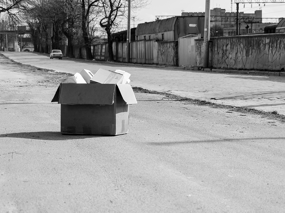 A black and white photo of a cardboard box filled with more boxes lies in the middle of the street.