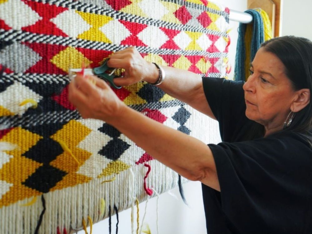Debra Sparrow, a middle-aged Musqueam woman with medium-light skin tone and straight dark hair, uses a pair of scissors to trim up some loose strands on a weaving that has been hung on a wall.