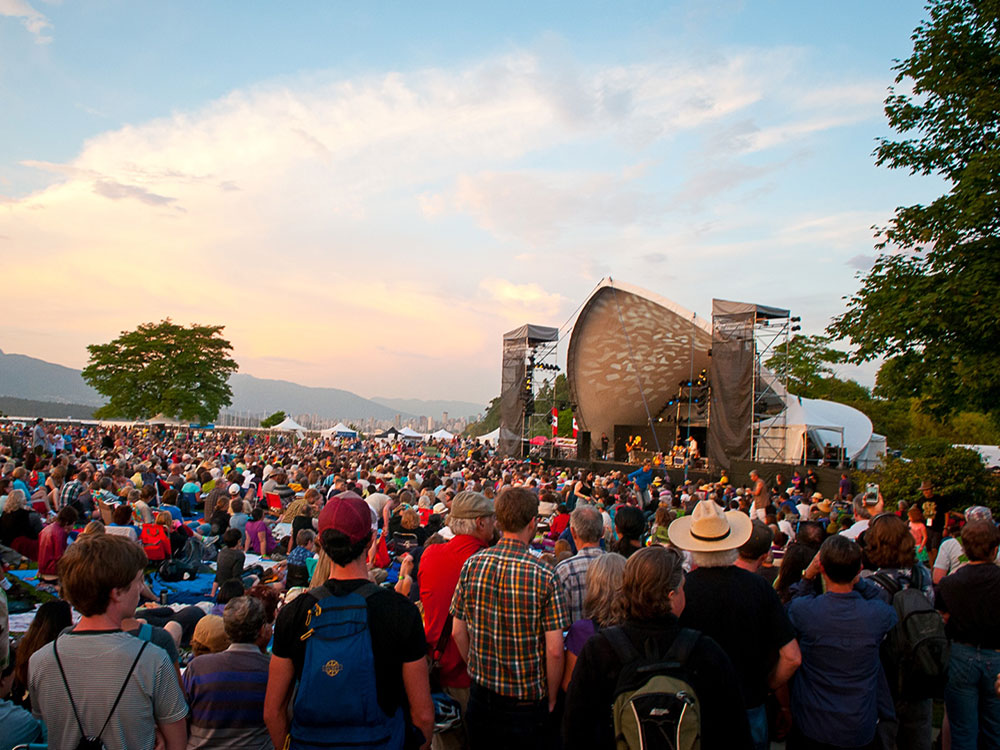 A large crown gathers around a performance stage at dusk, surrounded by mountains and party cloudy skies.