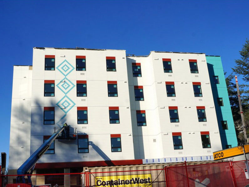 A white five-storey building under construction stands in front of a blue sky surrounded by a construction fence. A person is painting insignias on the wall via an aerial lift.