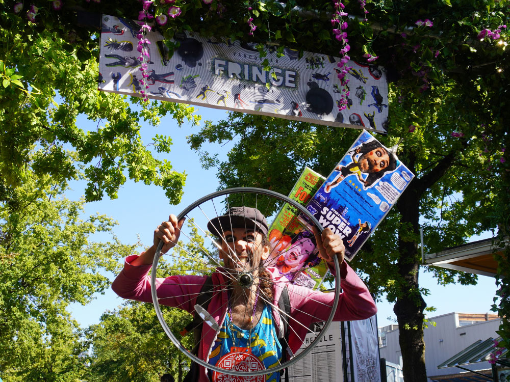 A person stands under a sign that reads ‘Vancouver Fringe’ hanging from a tree canopy. They are holding a bicycle wheel and grinning through it directly at the camera.
