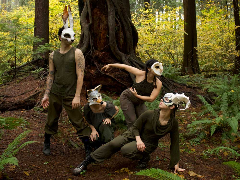 Four dancers dressed in earth tones and animal masks pose in the forest in front of a large tree trunk.