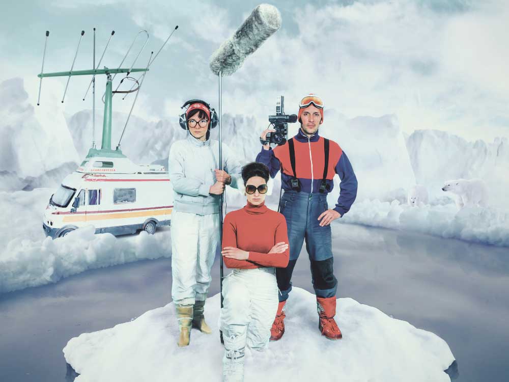 A staged photo of three people standing on a piece of floating ice. One is holding a video camera, another is holding a mic covered in a large, furry outdoor windscreen. A vintage van is behind them.
