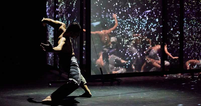 A man in loose pants dances shirtless in front of a reflective stage frame. A curtain of twinkling lights is behind the frame.