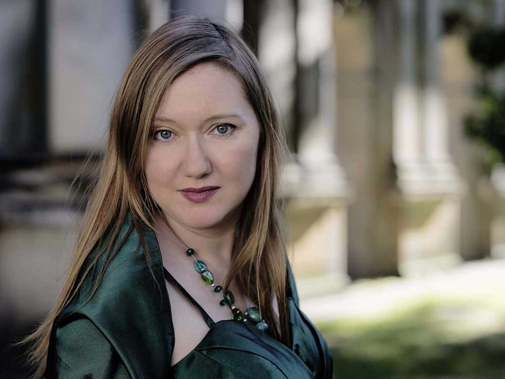 A light-skinned woman with long, straight blonde hair looks directly at the camera. She is wearing an elegant emerald green dress and shawl with matching necklace. Behing her is a blurred building with classic columns.